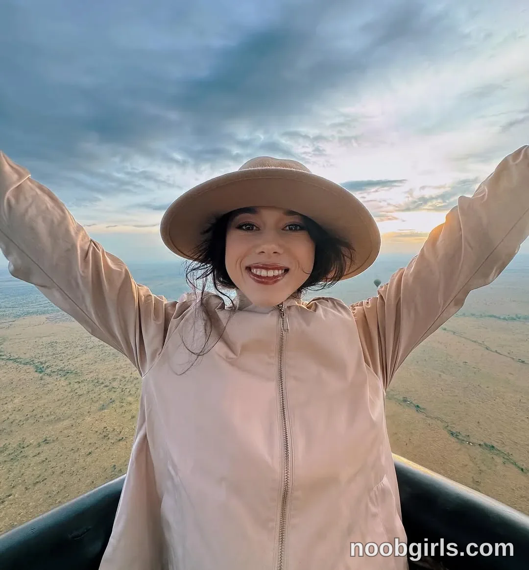 TAANNI facing the camera with both arms raised outward wearing light beige zip-front jacket in a basket above open grassland