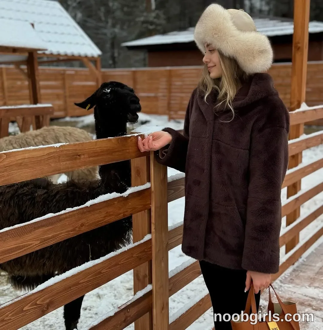 tastypoint standing beside a wooden fence wearing cream or light beige fur trapper-style hat in animal enclosure or farm pen