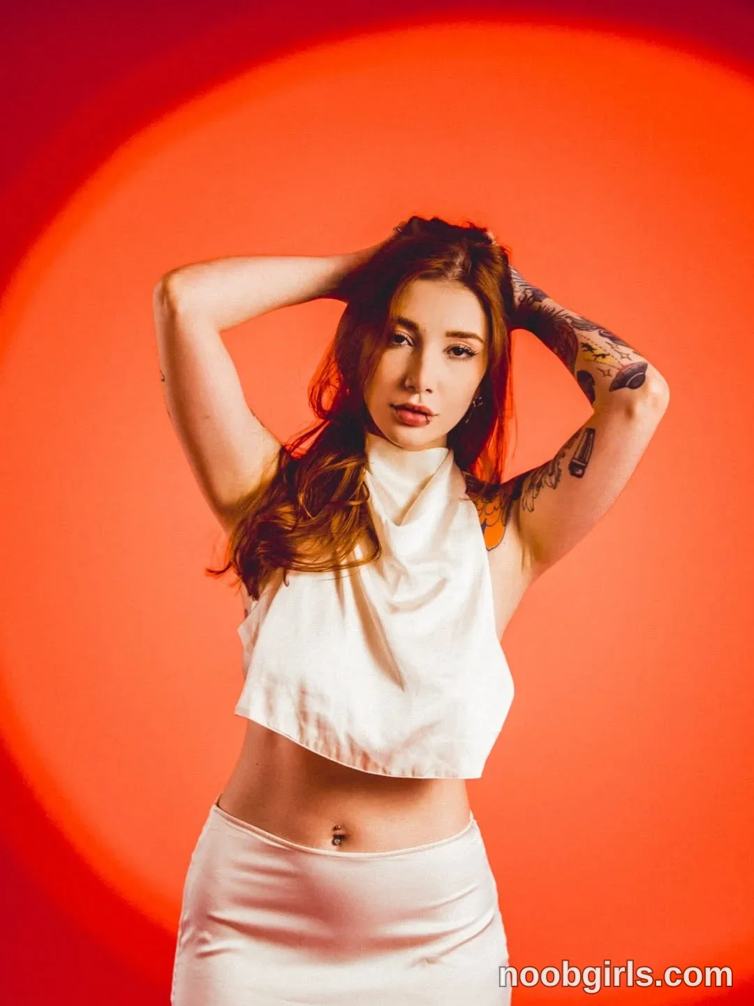 Alicia Standing facing the camera with both arms raised wearing White sleeveless cropped top with a draped neckline