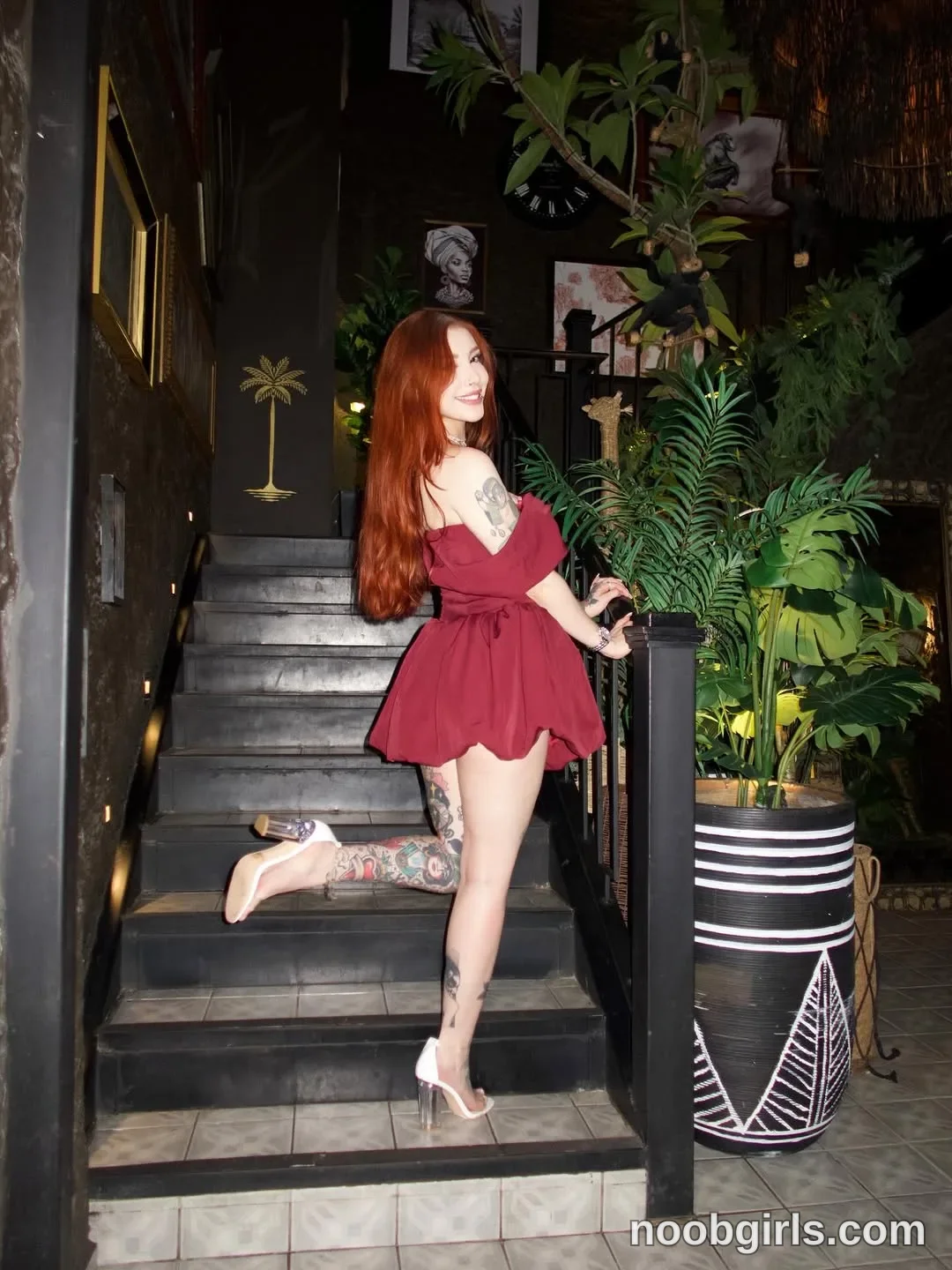 Alicia Standing sideways on the staircase with her hands resting on the railing wearing Burgundy off-the-shoulder short dress