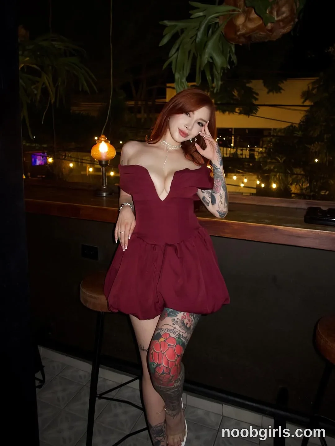 Alicia Standing with her legs crossed wearing Burgundy off-the-shoulder mini dress with a plunging neckline and puffed skirt
