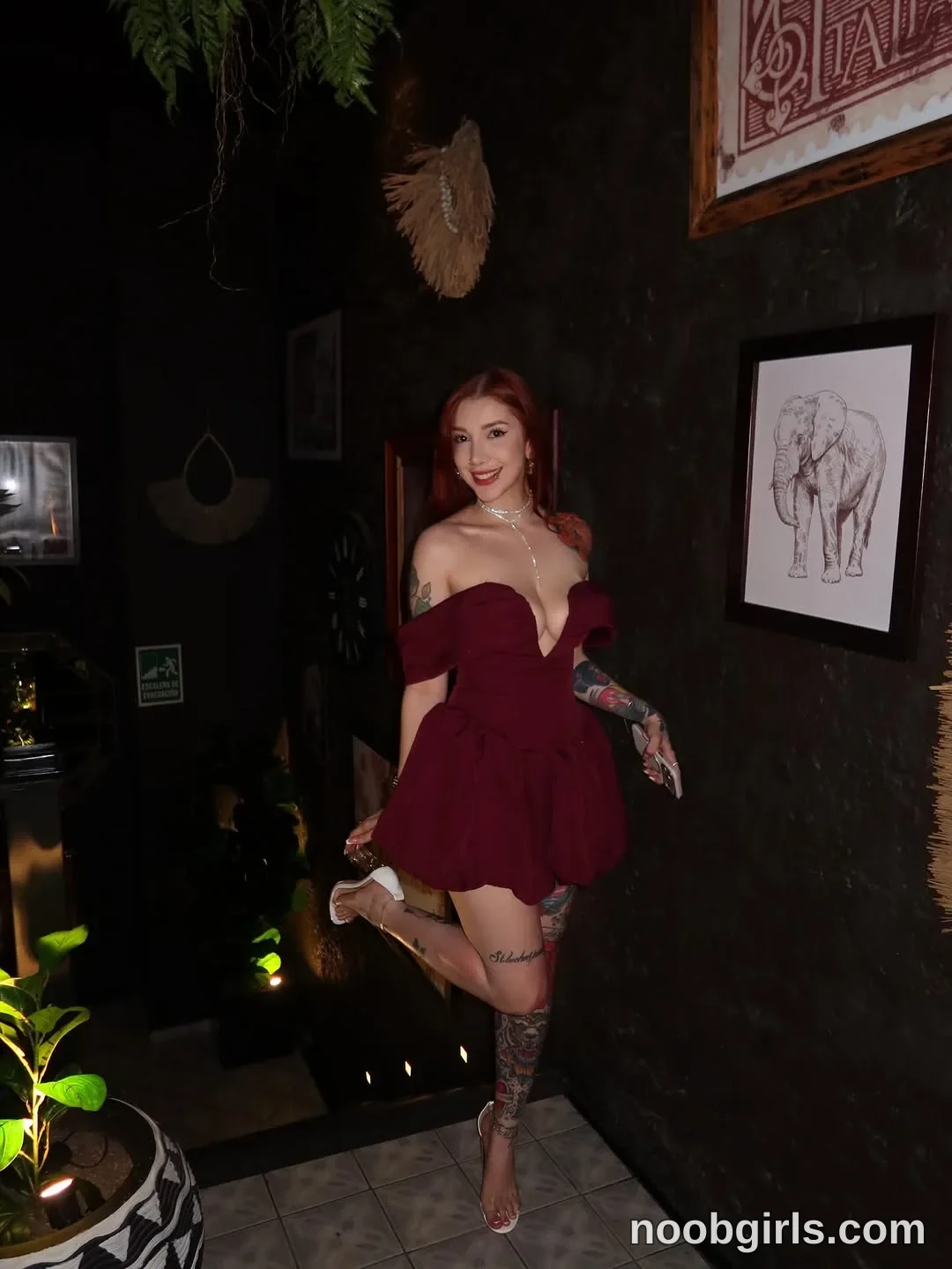 Alicia Standing on one leg with the other knee bent wearing Burgundy off-the-shoulder short dress with a plunging neckline