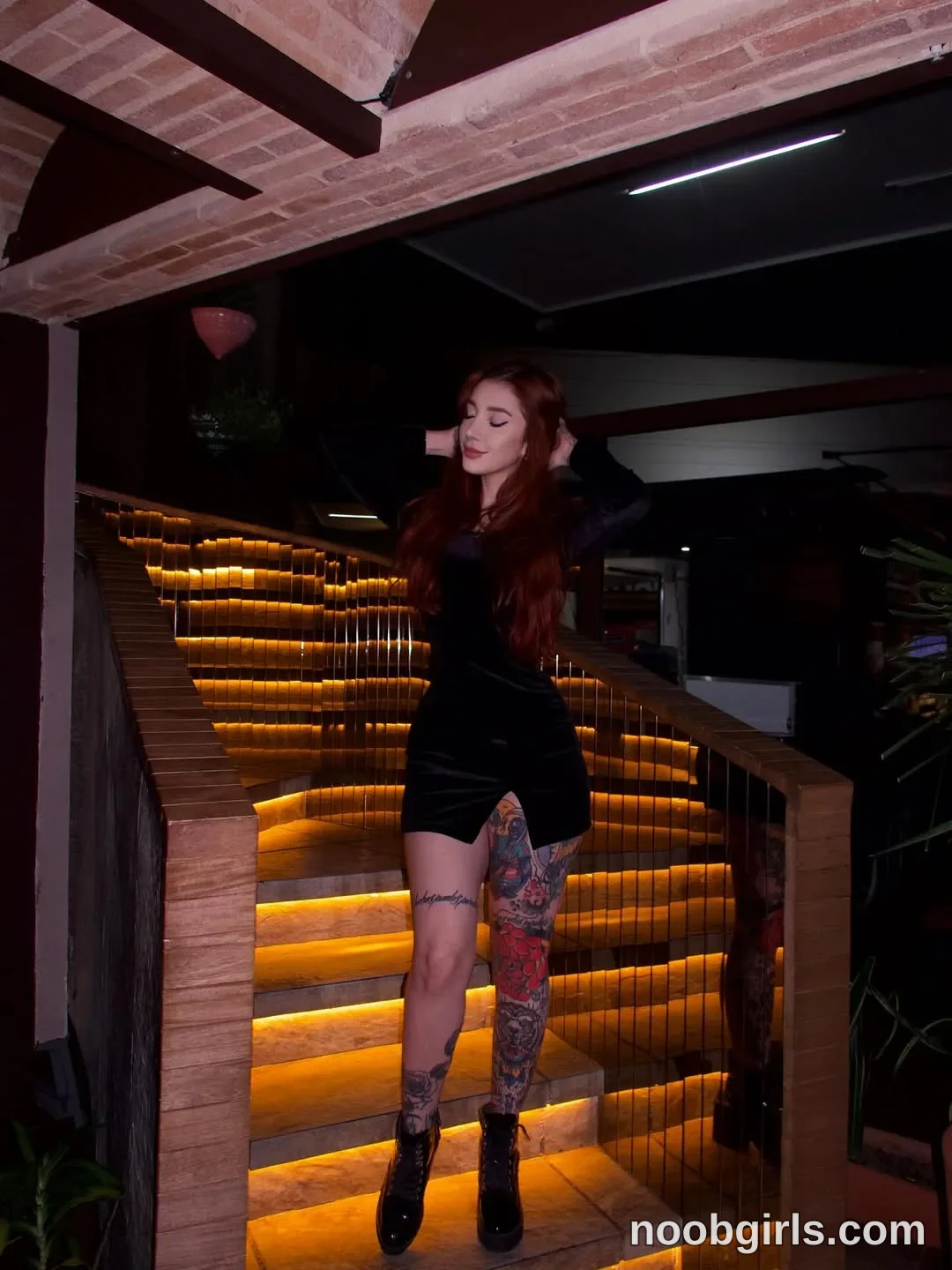 Alicia Standing on the stairs with both arms raised wearing Black long-sleeved mini dress with a slit at the front hem