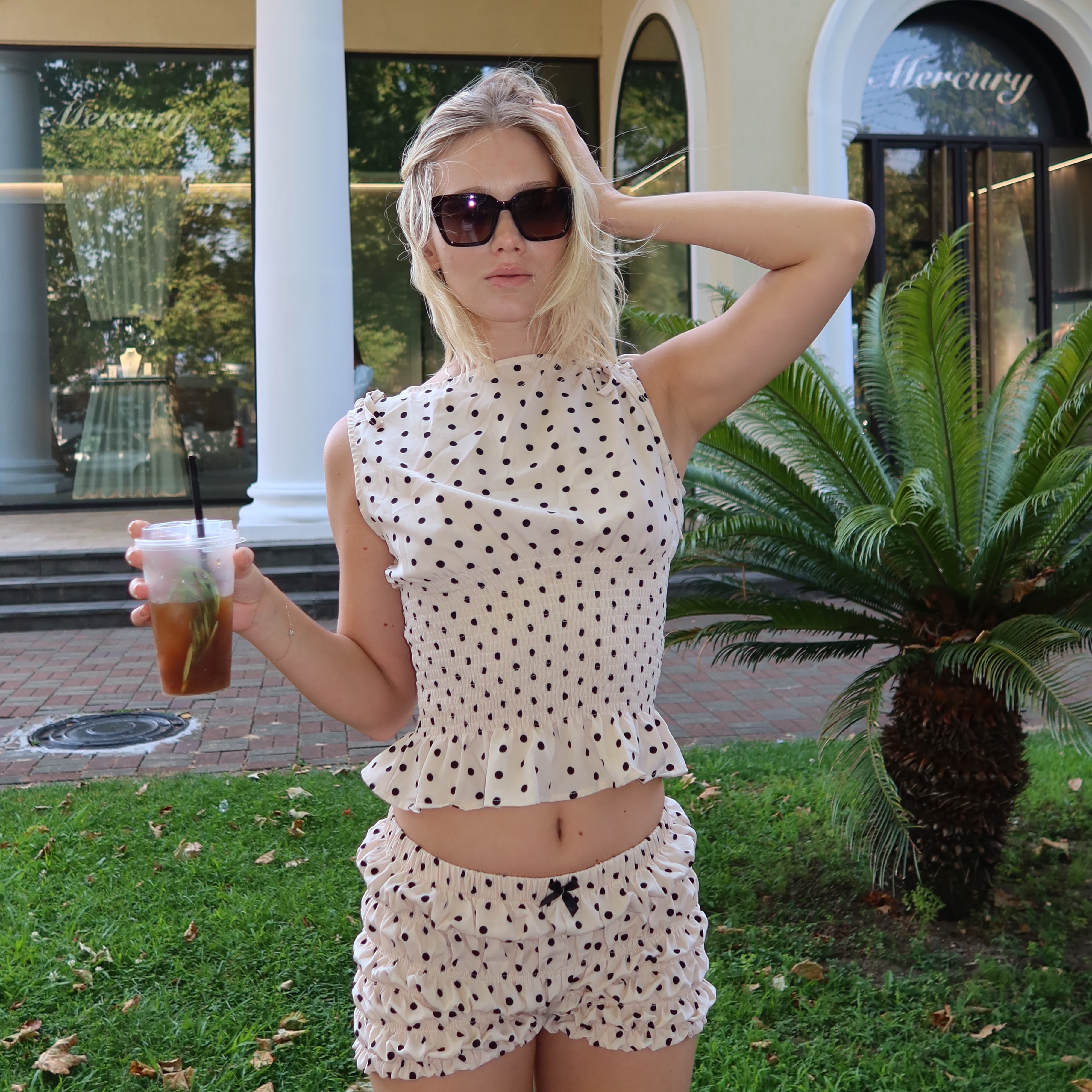 tollysage holding an iced beverage wearing cream polka-dot sleeveless top in paved walkway