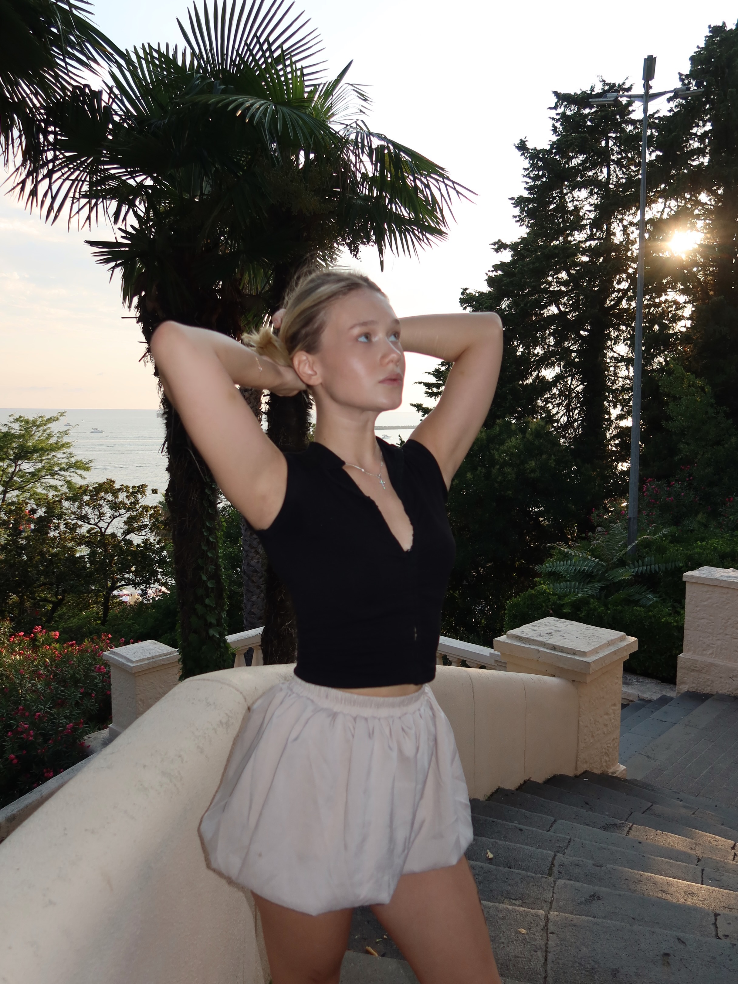 tollysage holding hair with both hands wearing black sleeveless crop top in stone staircase overlooking sea