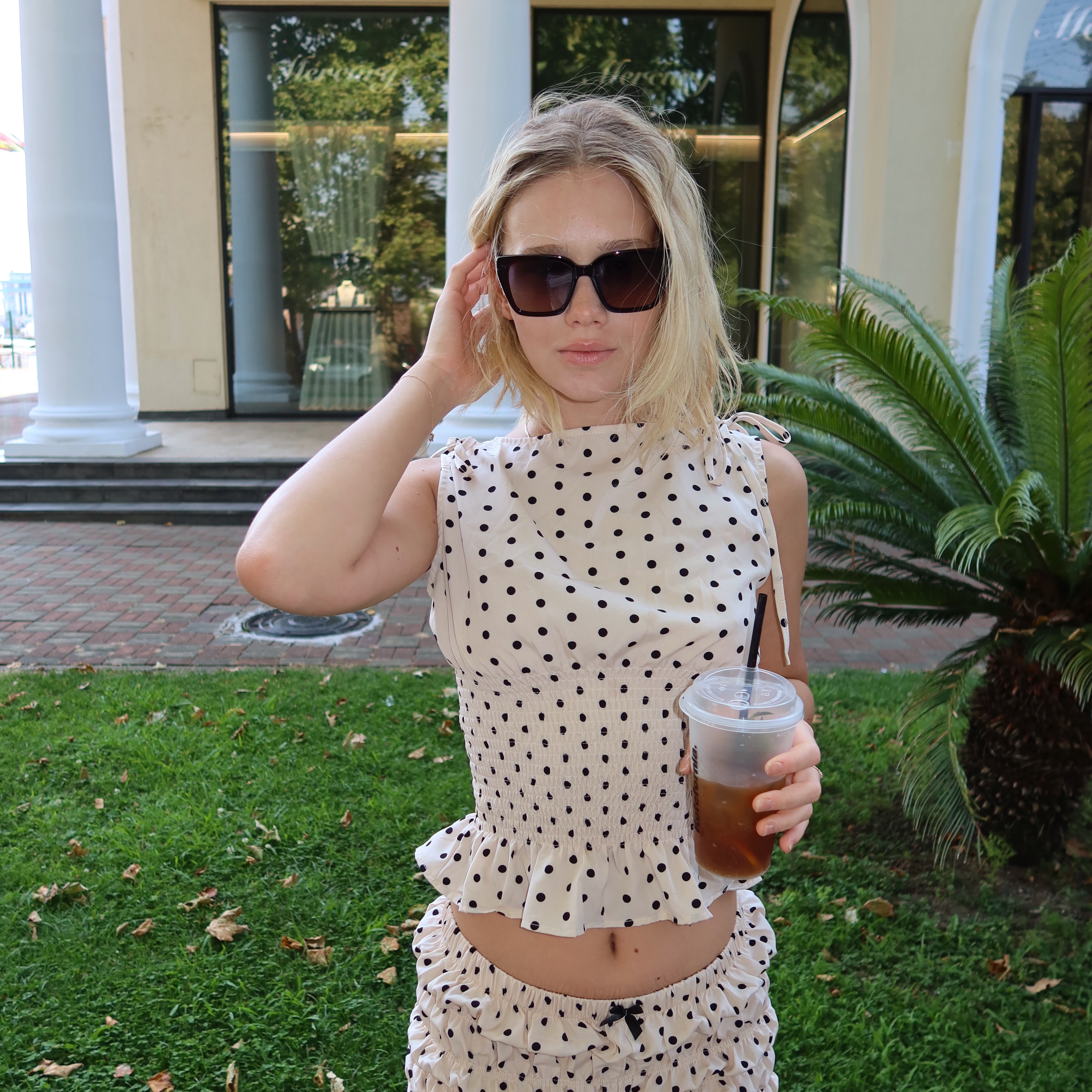 tollysage holding an iced drink wearing white polka dot top in lawn