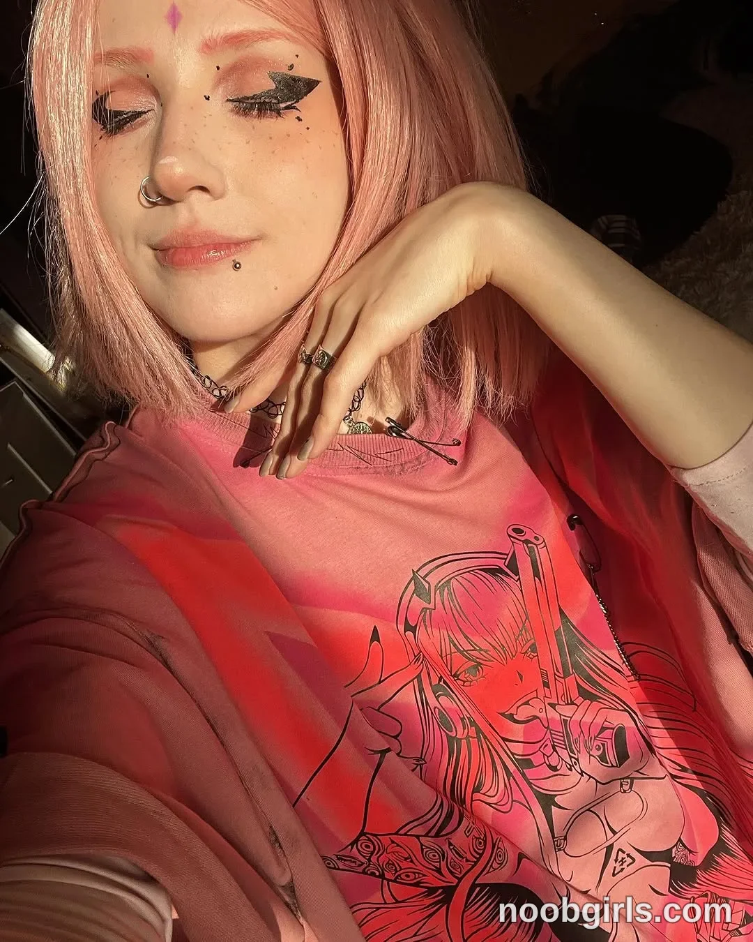 Lina seated with one hand raised beside her face wearing pink graphic T-shirt in likely inside a vehicle