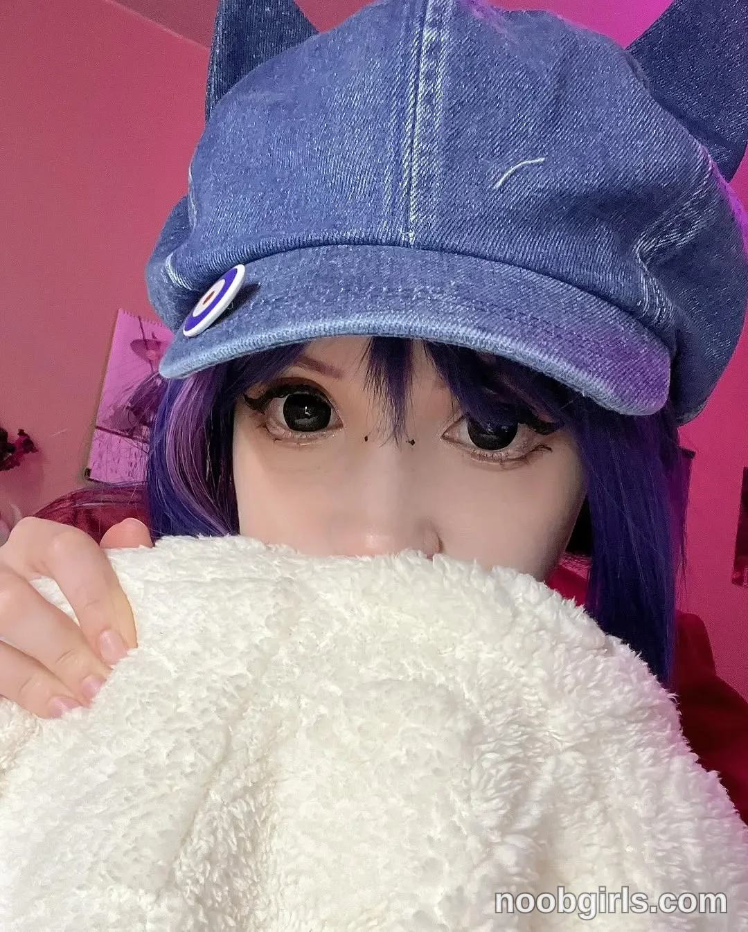 Lina holding a white fluffy blanket up over the lower face wearing blue denim cap in room
